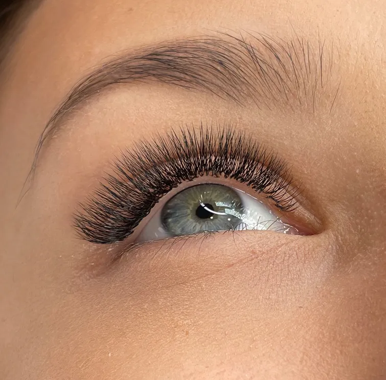 Close-up of a blue-green eye with dense, curled volume lash extensions and a natural eyebrow