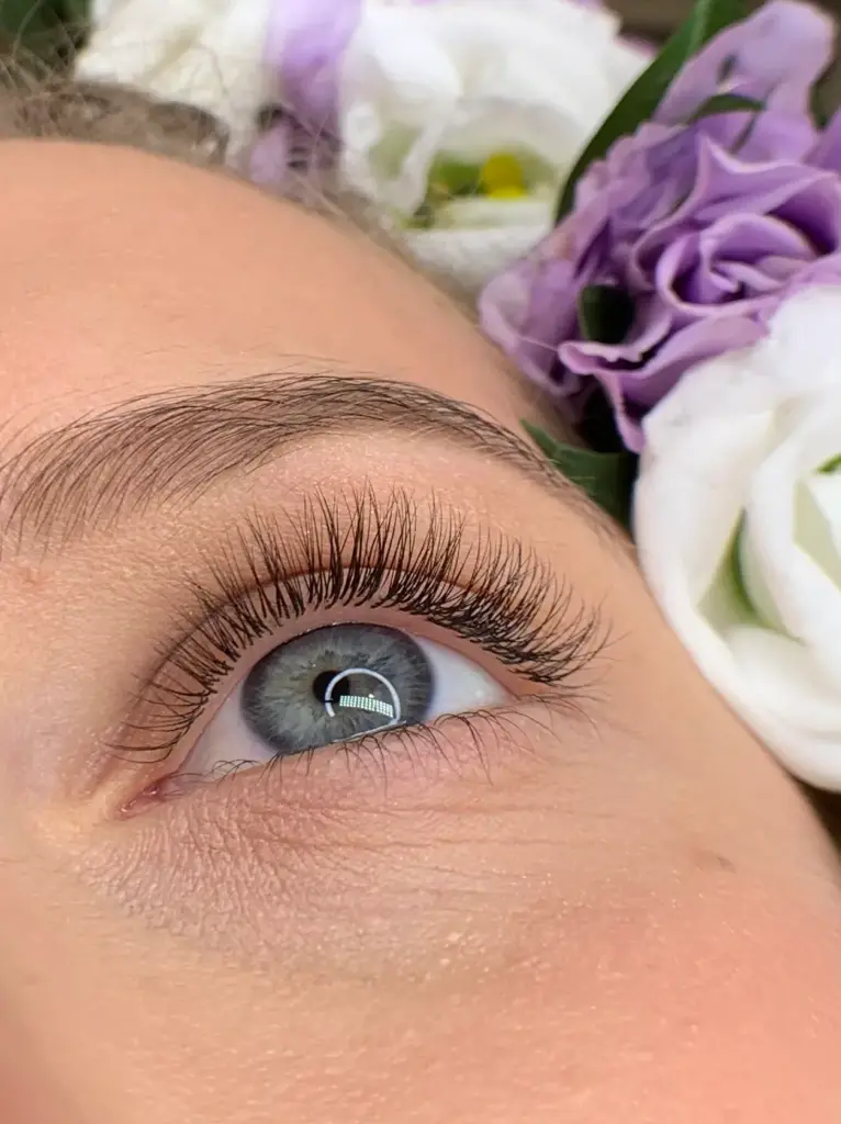 Close-up of a blue-gray eye with long classic lash extensions and a shaped brow beside purple and white flowers.