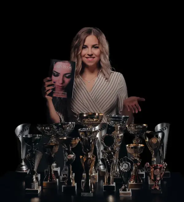 Alexa Borbély holds a book titled “Lash Book” behind a table crowded with trophies against a black background.