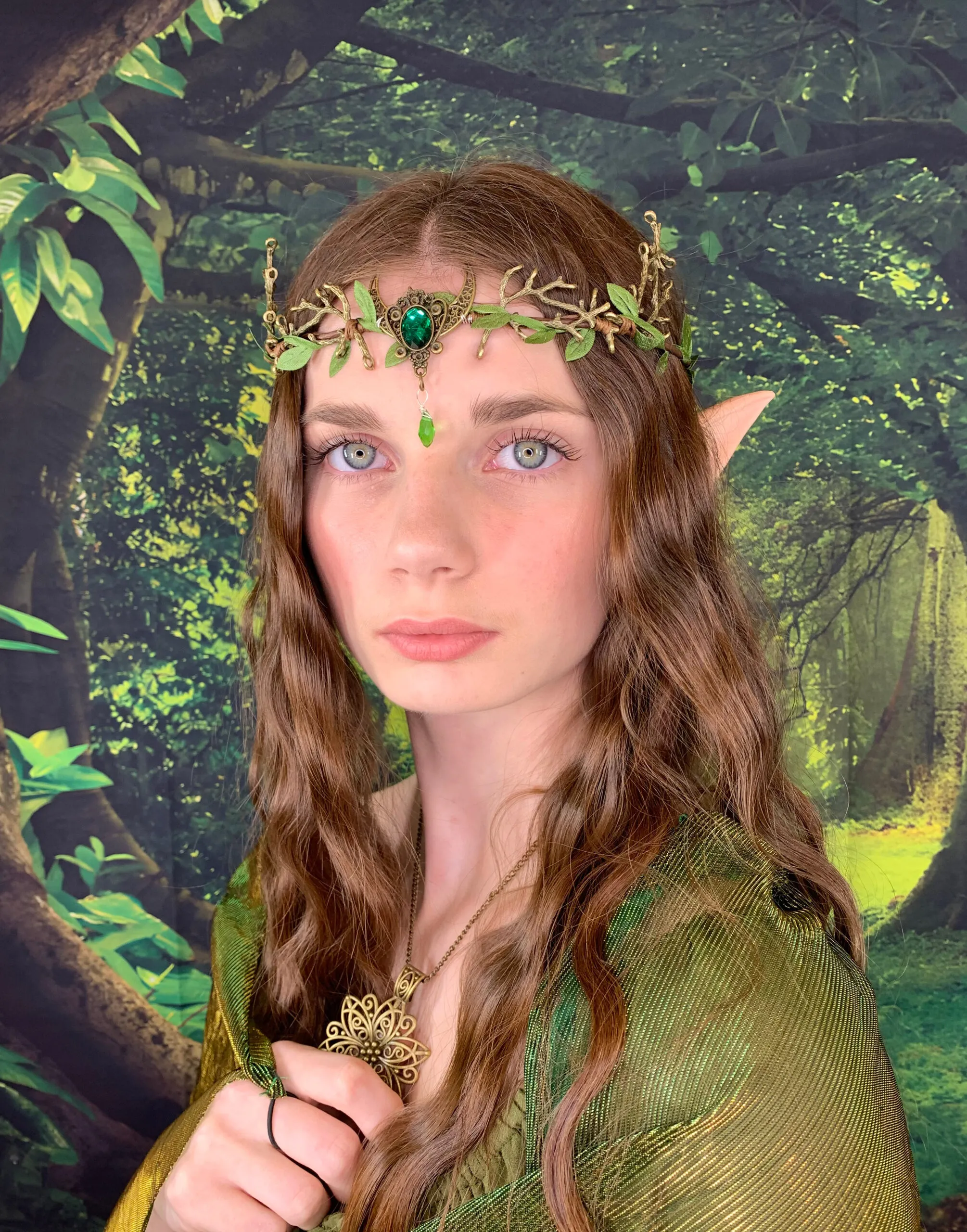 Model in fantasy elf styling with long lashes, pointed ears, a green jeweled headpiece and forest backdrop.
