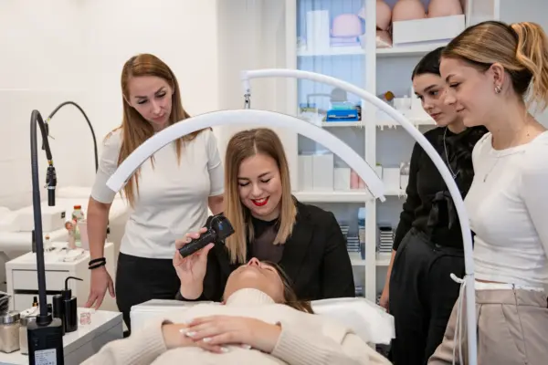 An instructor demonstrates lash extension work on a reclining client as trainees observe in a beauty studio.