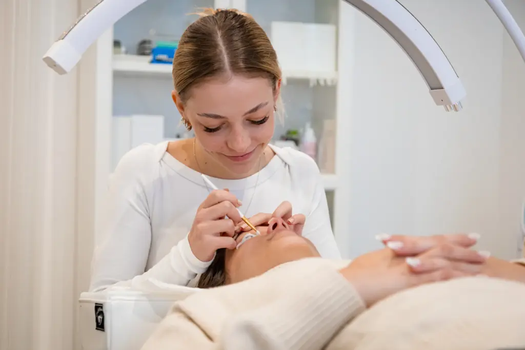 A lash artist uses a fine tool to style a reclining client's eyelashes in a bright salon.
