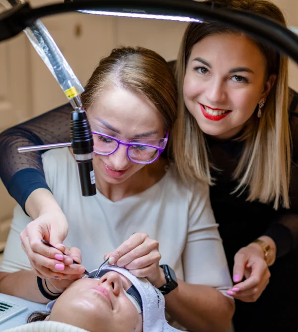 A lash technician applies extensions to a reclining client under a UV/LED lamp as an instructor observes.