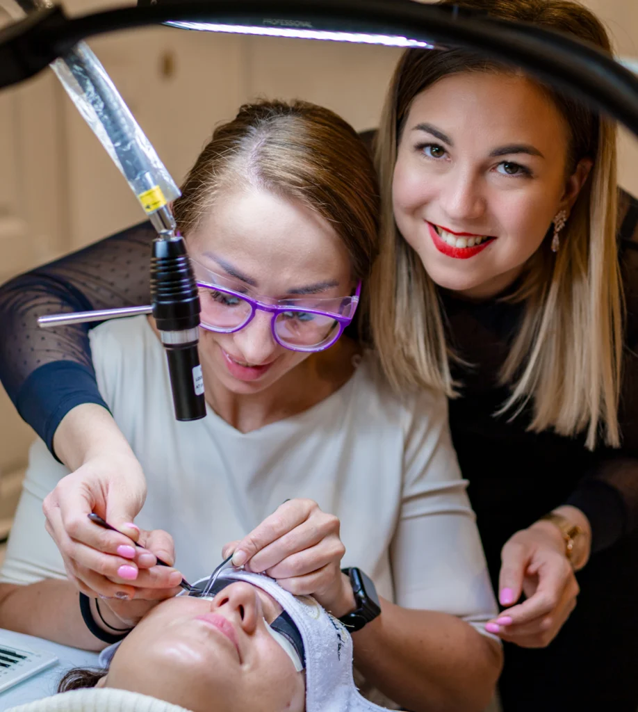 A lash technician applies extensions to a reclining client under a UV/LED lamp as an instructor observes.