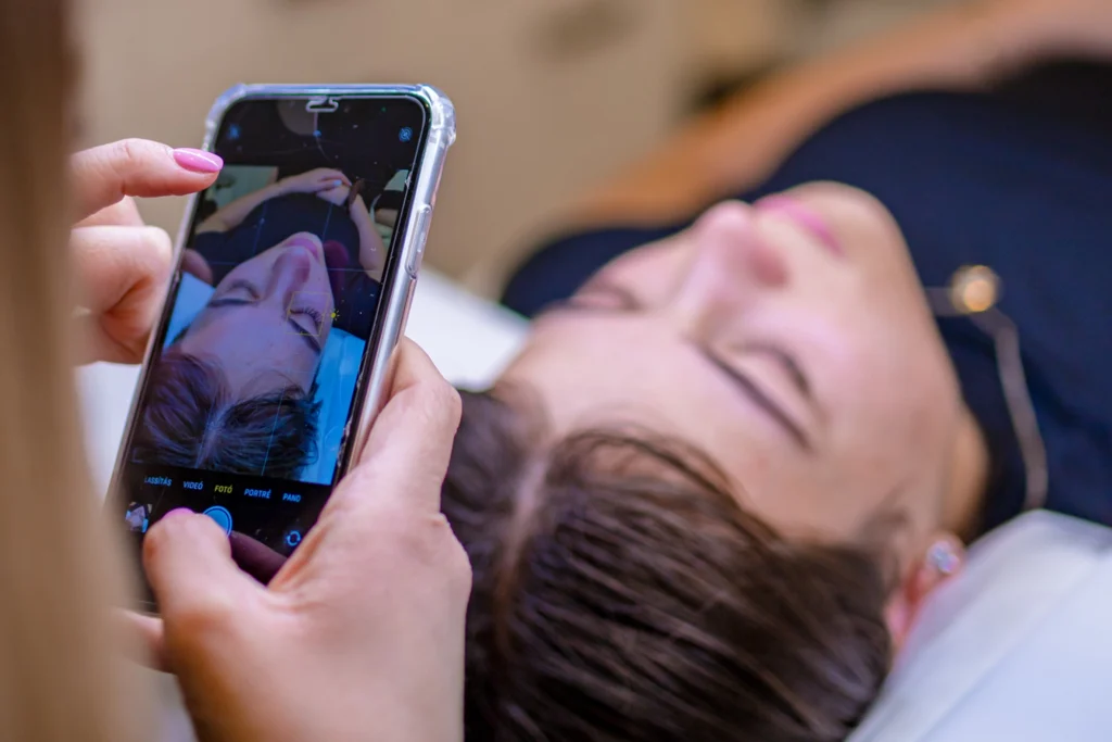 A stylist photographs a reclining woman’s closed eye and eyelashes with a smartphone.