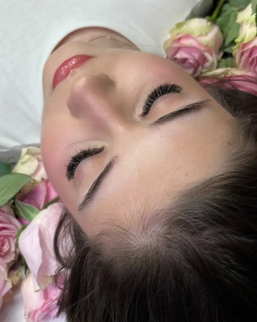 Close-up of a woman with voluminous eyelash extensions and groomed brows, lying among pink roses.