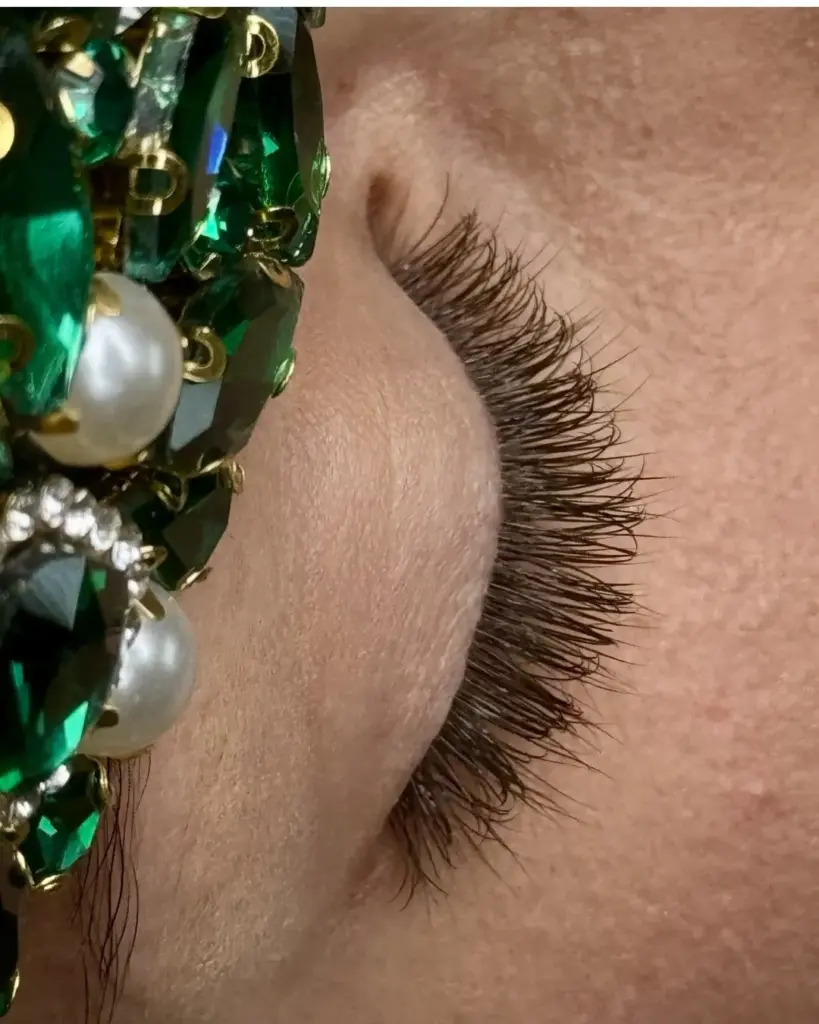 Close-up of long, curled dark eyelash extensions beside a green jeweled hair accessory