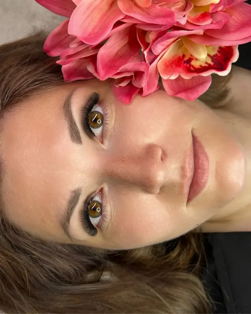 Close-up portrait of a woman with long, full lashes and shaped brows beneath pink flowers.