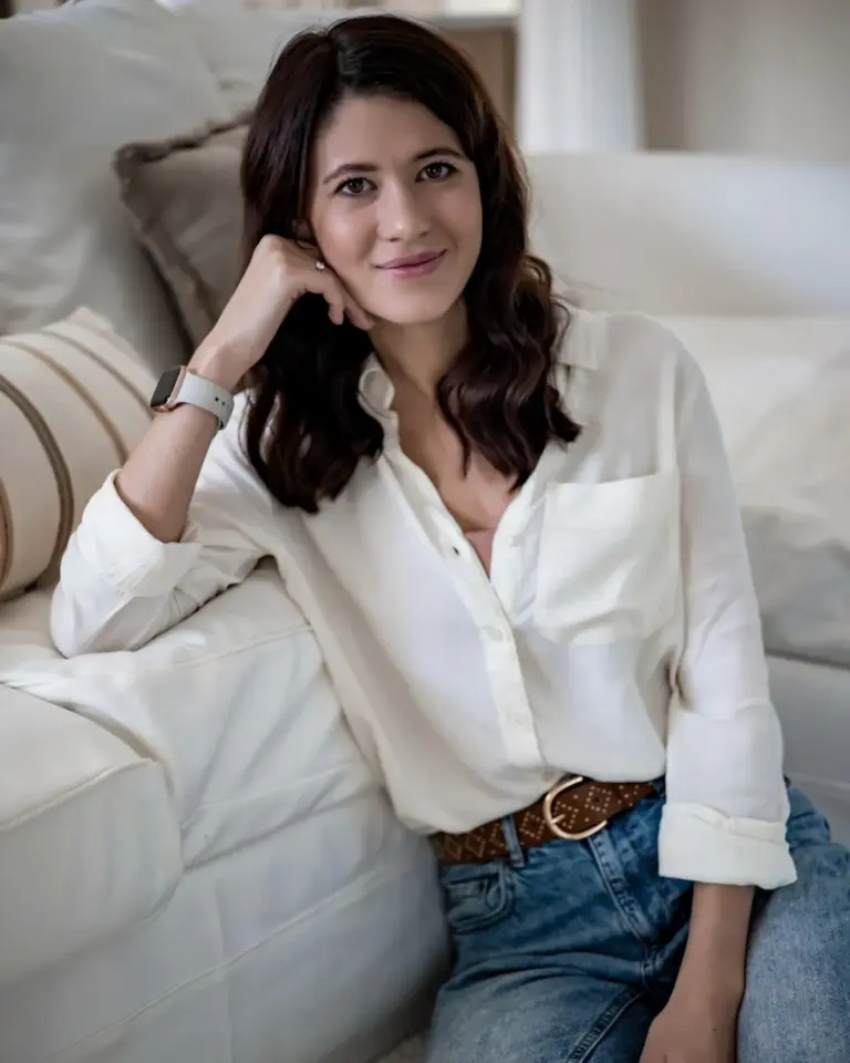 Kata Hajnal-Ligeti smiles while seated on a white sofa, wearing a white blouse, jeans, and a smartwatch.