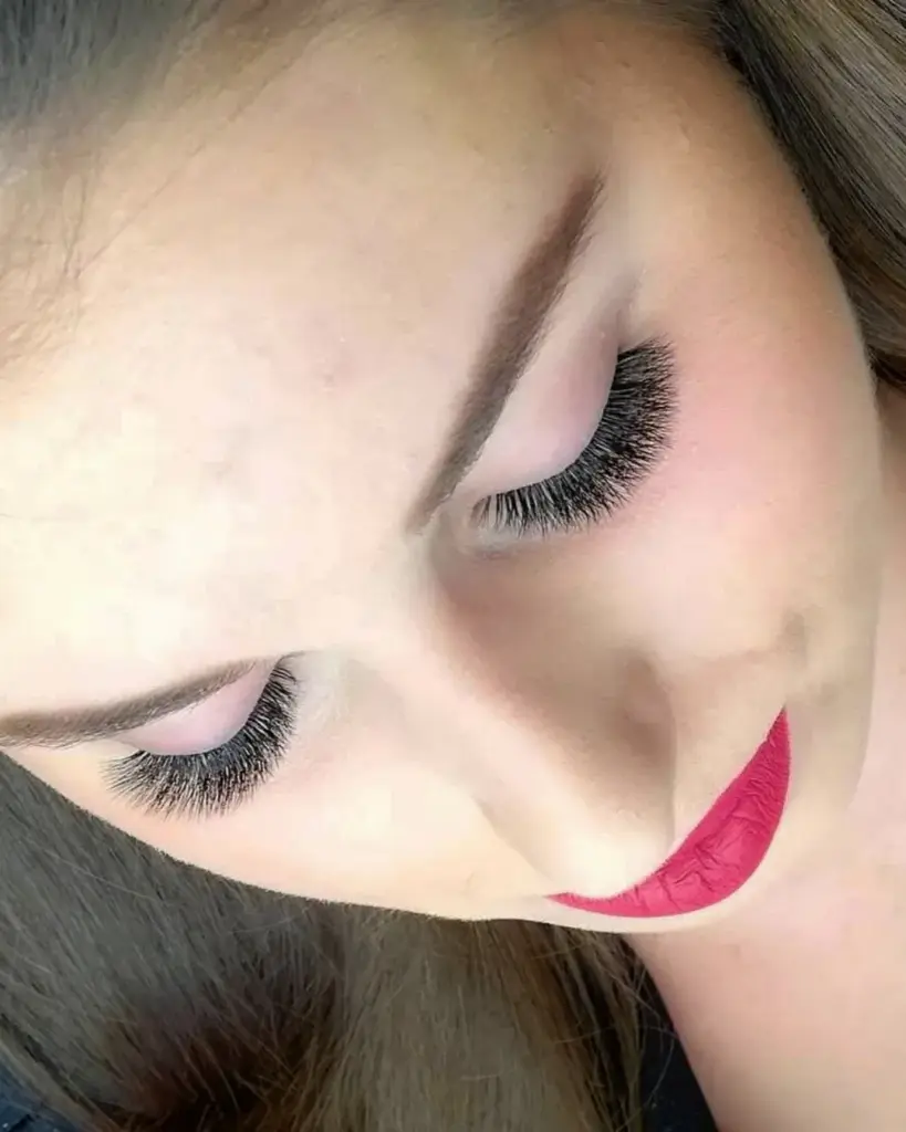 Close-up of a woman with closed eyes, voluminous lash extensions, shaped brows, and bright pink lipstick.