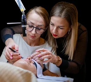 Two women practice eyelash styling on a reclining client, carefully applying extensions with tweezers.