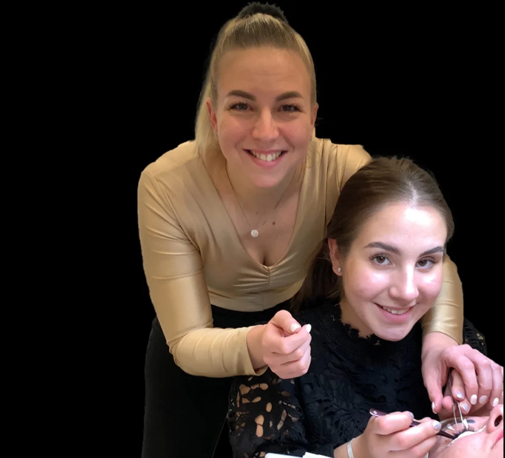 Lash trainer guides a seated student as she applies extensions to a reclining client.