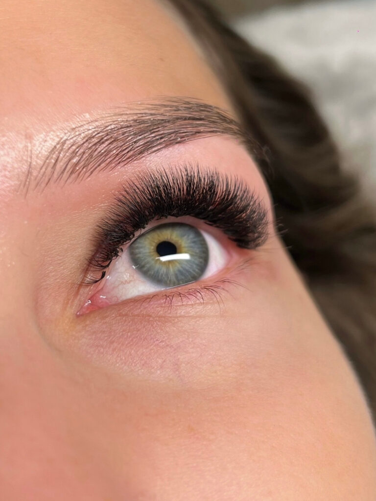 Close-up of a green eye with full, curled lash extensions and a neatly shaped eyebrow.