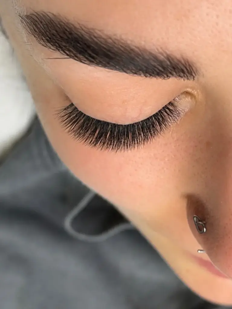 Close-up of a closed eye with dense, curled lash extensions, a shaped eyebrow, and a nose piercing.
