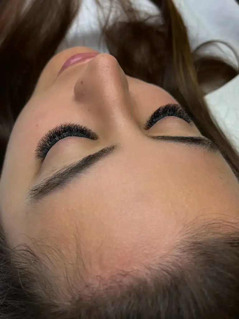 Close-up of a reclining client with closed eyes, full black lash extensions, and neatly shaped eyebrows.