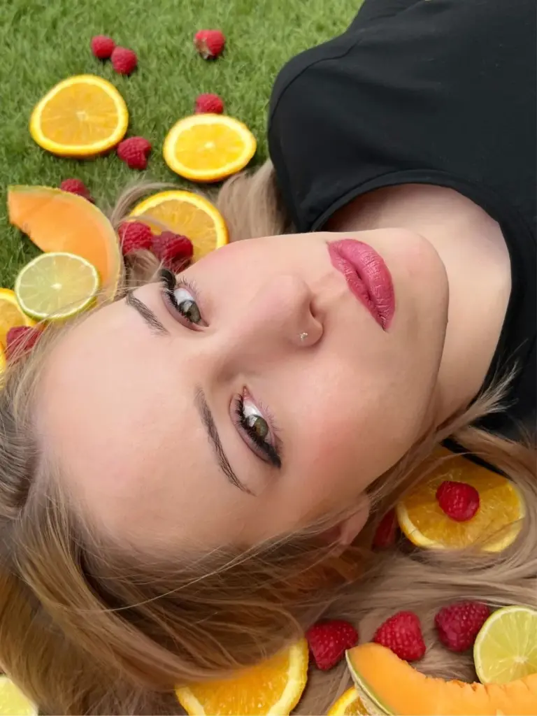 Close-up of a woman with defined brows, long lashes and pink lipstick lying among citrus slices and raspberries
