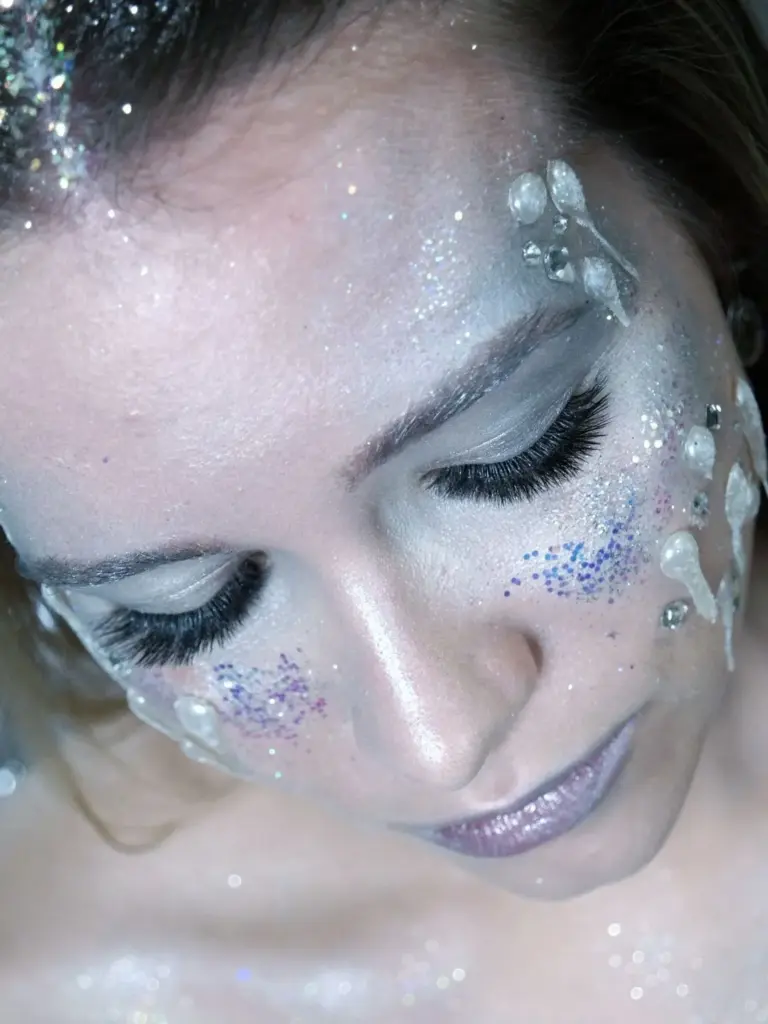 Close-up of a woman with closed eyes, long lashes, silver glitter makeup, and decorative gems