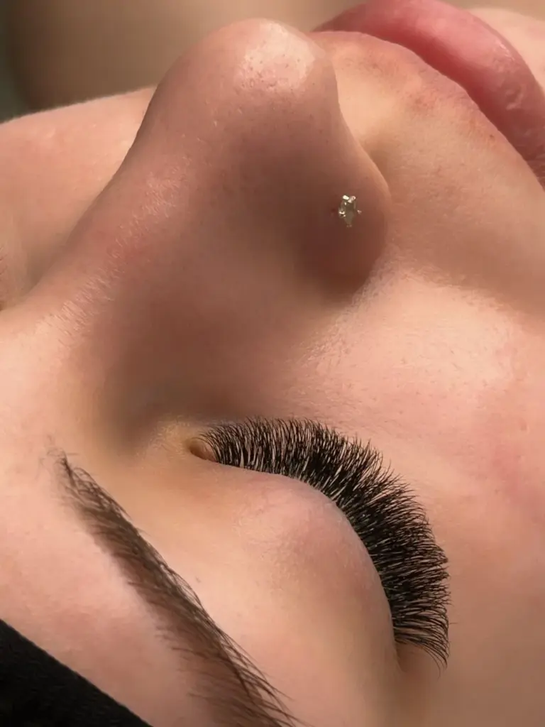 Close-up of a closed eye with dense, curled eyelash extensions, a shaped brow, and a nose stud.
