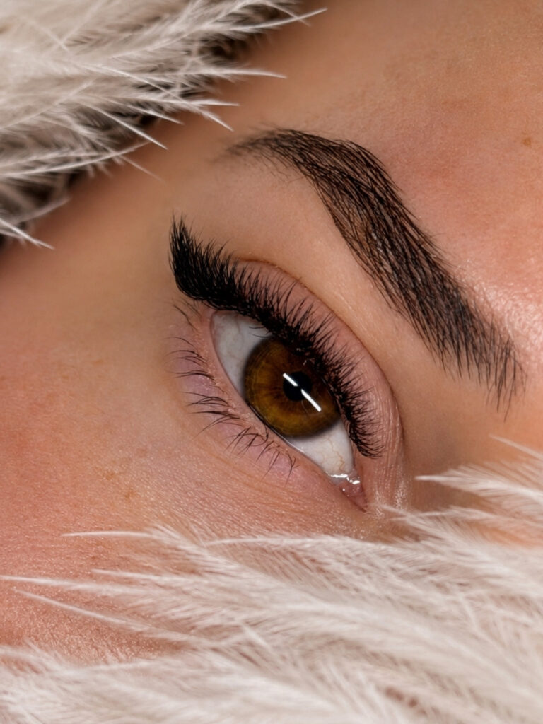Close-up of an amber eye with long curled lashes and a defined dark brow, framed by soft white feathers.