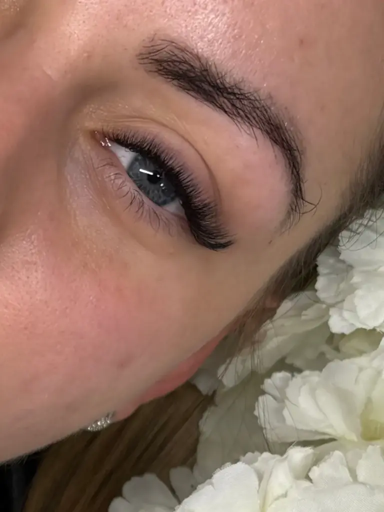 Close-up of a blue-gray eye with long, curled lash extensions and a defined eyebrow beside white flowers.