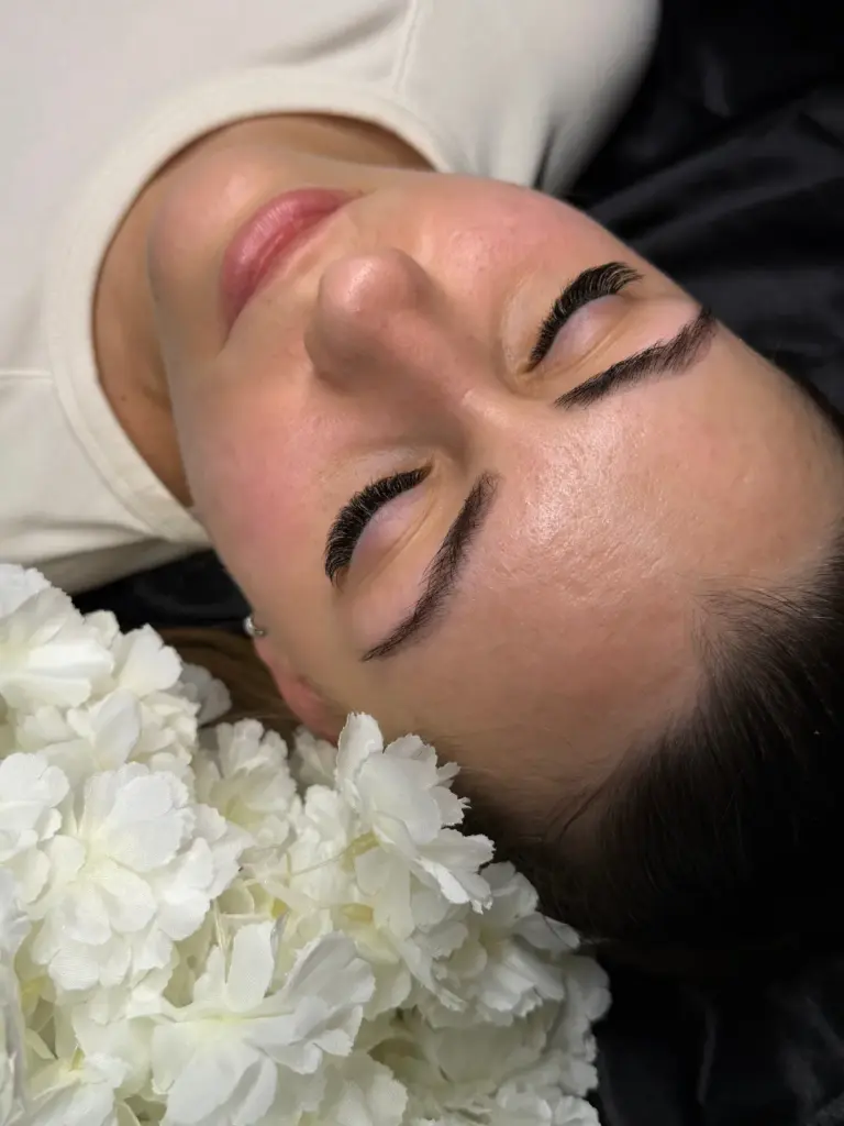 Woman with eyes closed, showing long lash extensions and defined brows beside white flowers.