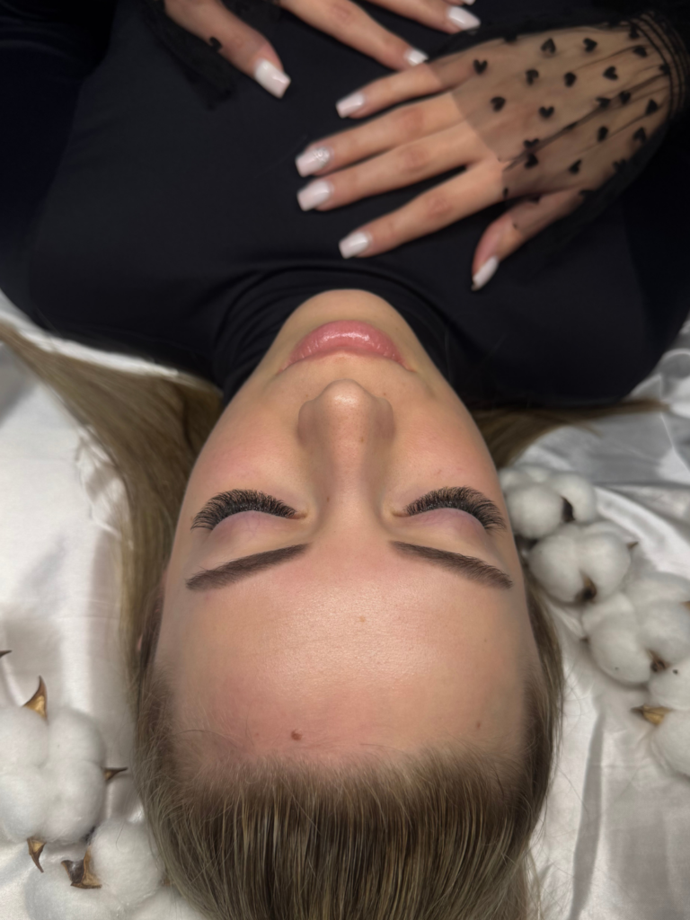 Woman lying with eyes closed, showing full eyelash extensions and shaped brows on a cotton-decorated beauty bed