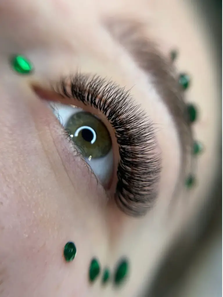 Close-up of a green eye with long, dense black lash extensions and green decorative accents on the skin.