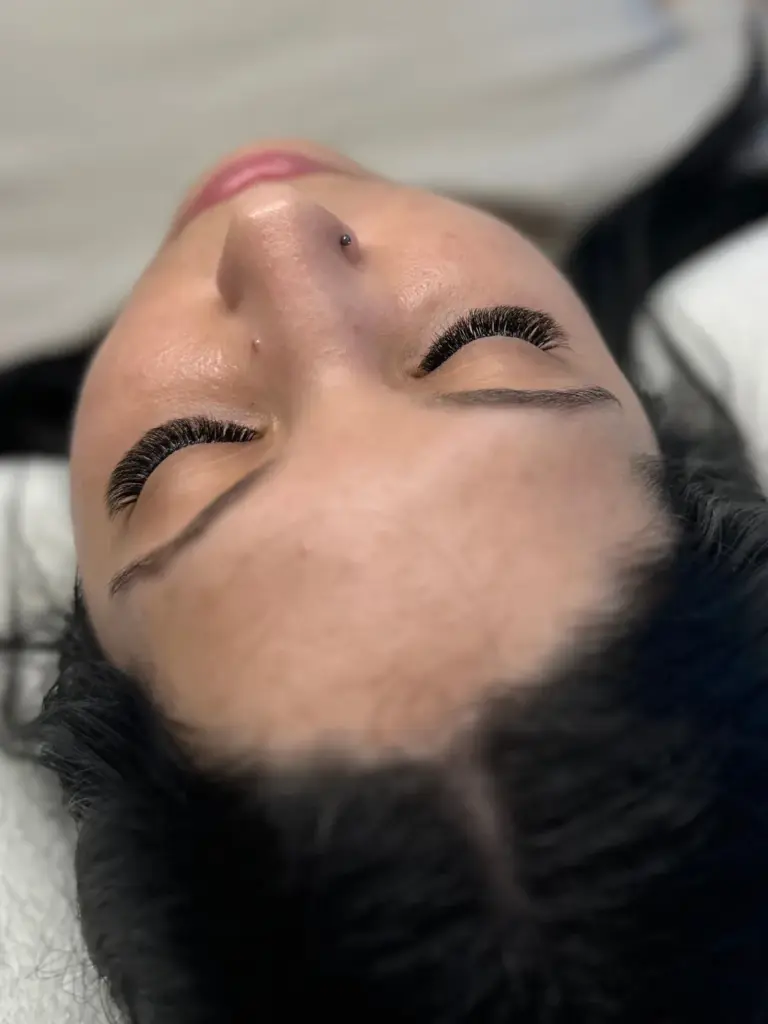 Close-up of a reclining woman with voluminous eyelash extensions, shaped brows, and a nose stud.