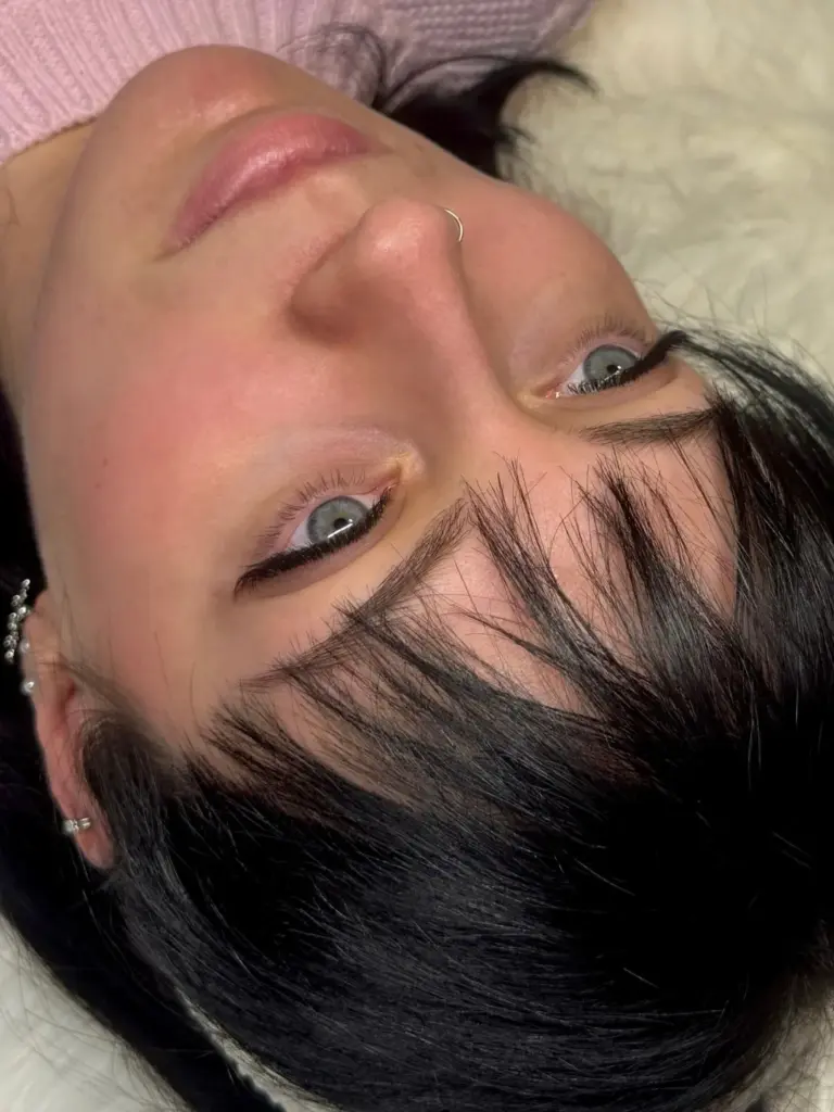 Close-up of a reclining woman with defined dark eyeliner, long lashes, natural brows, and a nose ring.