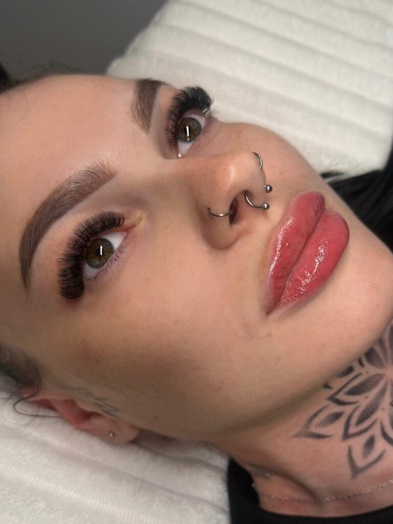 Close-up of a reclining client with voluminous lash extensions, shaped brows, glossy lips, and nose piercings.
