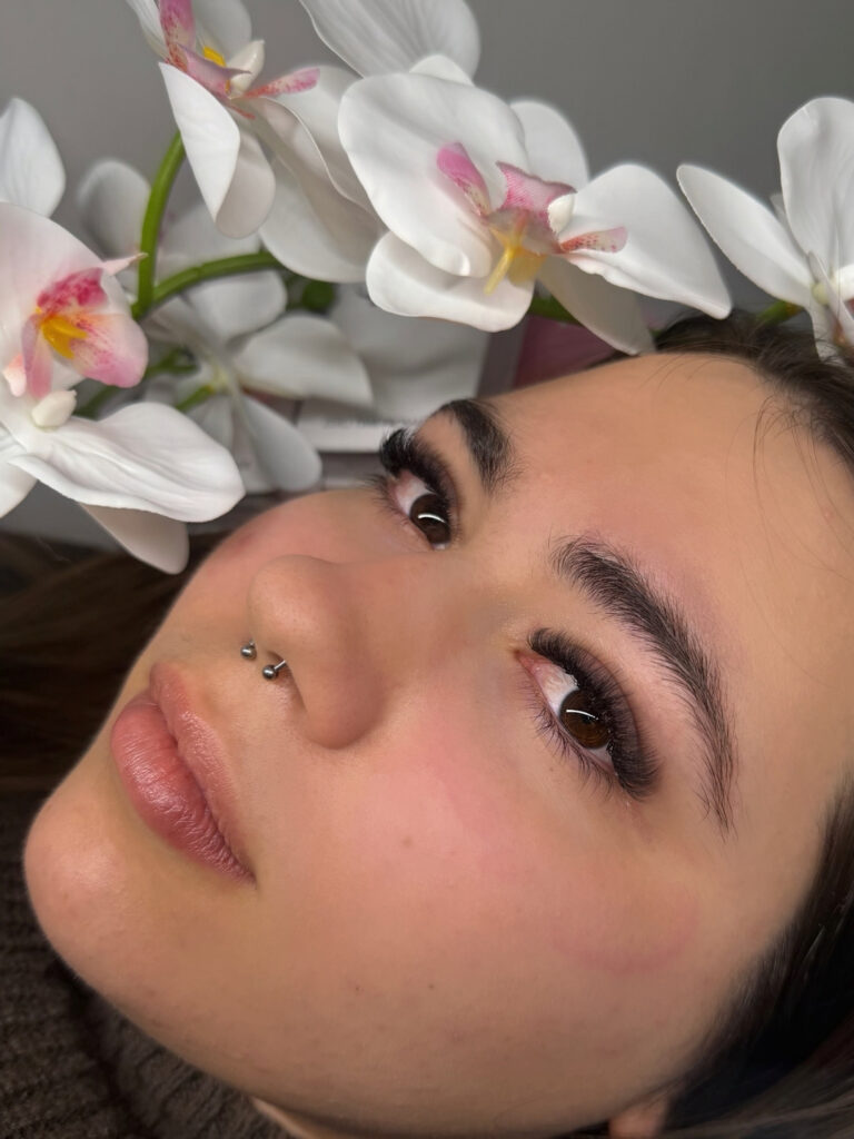 Close-up of a woman showcasing voluminous lash extensions and groomed brows beside white orchids.