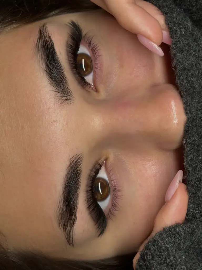 Close-up of a client with sculpted brows and long, voluminous lash extensions, framed by manicured hands.