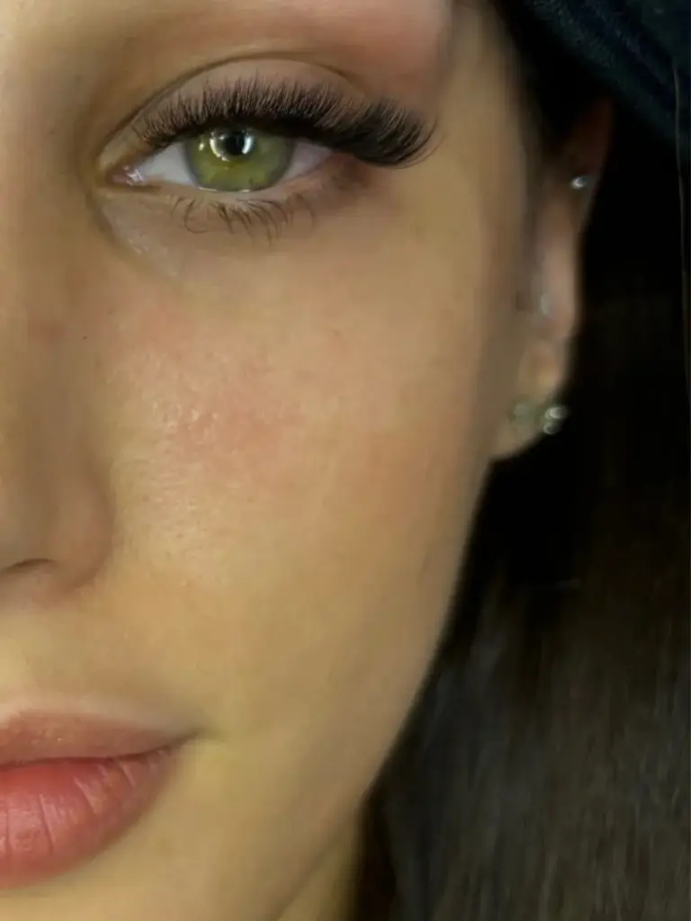 Close-up of a green eye with long, voluminous lash extensions, dark hair, and small ear piercings.
