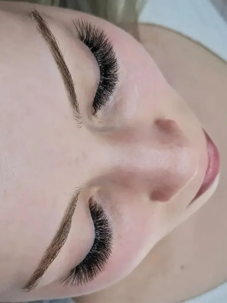 Close-up of a woman with closed eyes, full black eyelash extensions, and neatly shaped eyebrows.