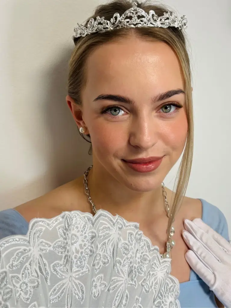Woman in a blue dress and jeweled tiara poses with a lace fan, showing styled lashes and brows.
