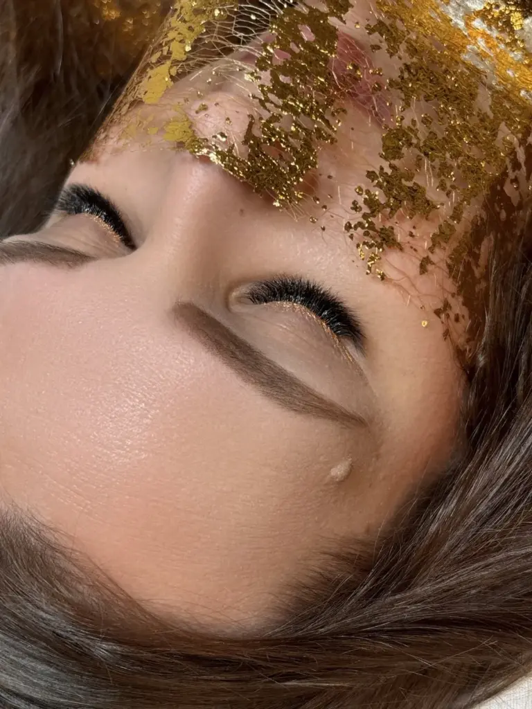 Close-up of a woman with long eyelash extensions, shaped brows, and gold leaf across her forehead.