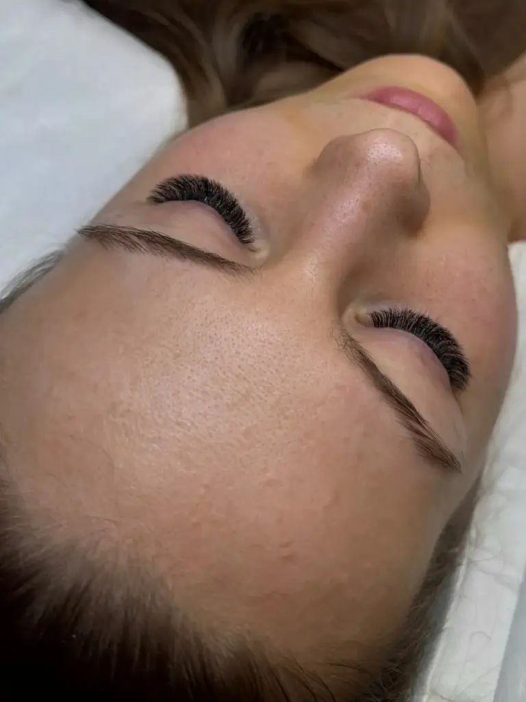Close-up of a woman lying with her eyes closed, showing thick lash extensions and neatly shaped brows.