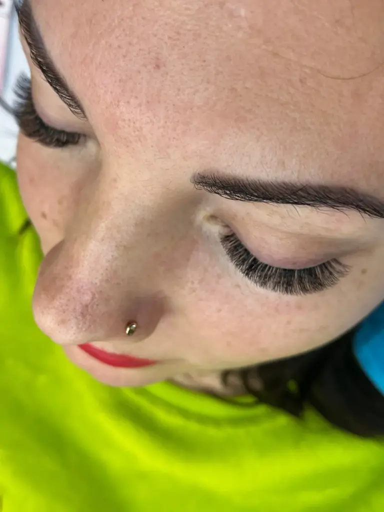 Close-up of a woman with eyes closed, showcasing long, voluminous eyelash extensions and a nose stud.