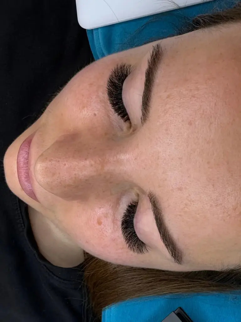 Close-up of a woman with eyes closed, wearing full lash extensions and neatly shaped brows on a salon bed.