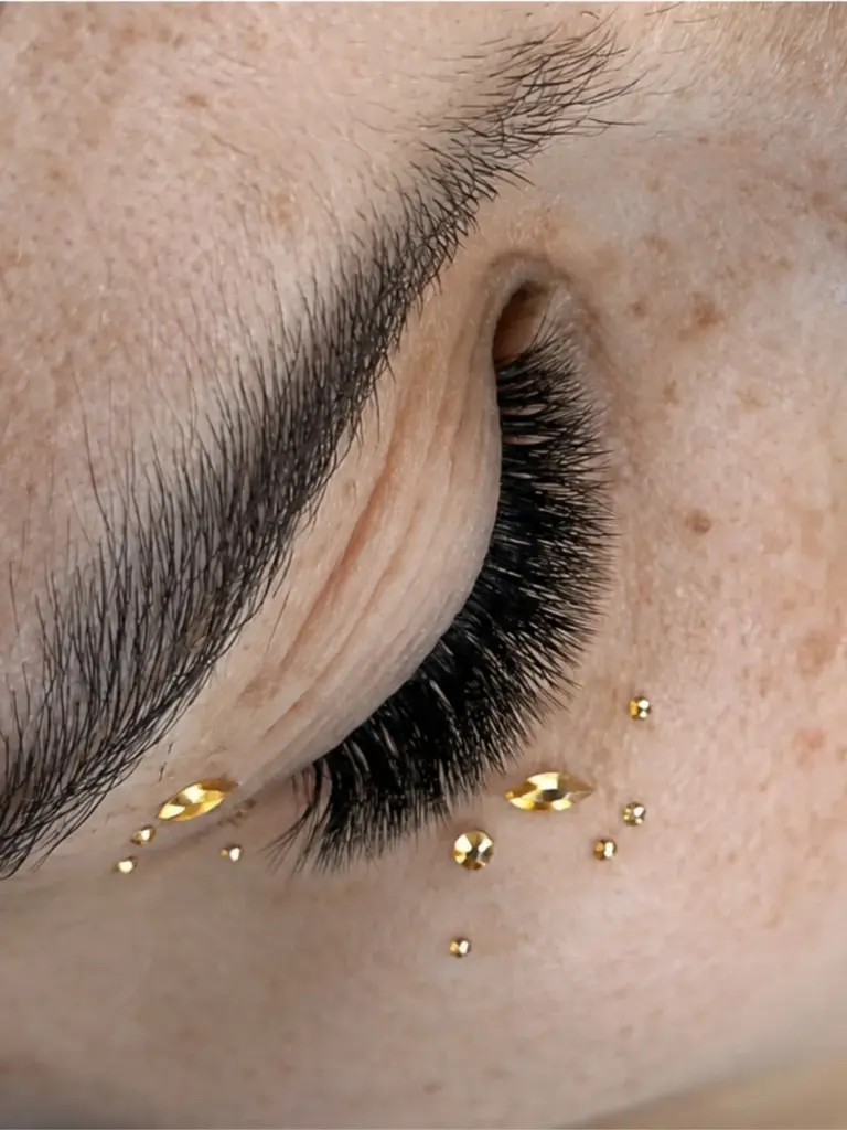 Close-up of a closed eye with long dark lash extensions, a shaped eyebrow, and gold facial gems