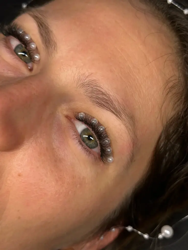 Close-up of a woman’s eyes with long curled lash extensions accented by small pearl beads and natural brows.