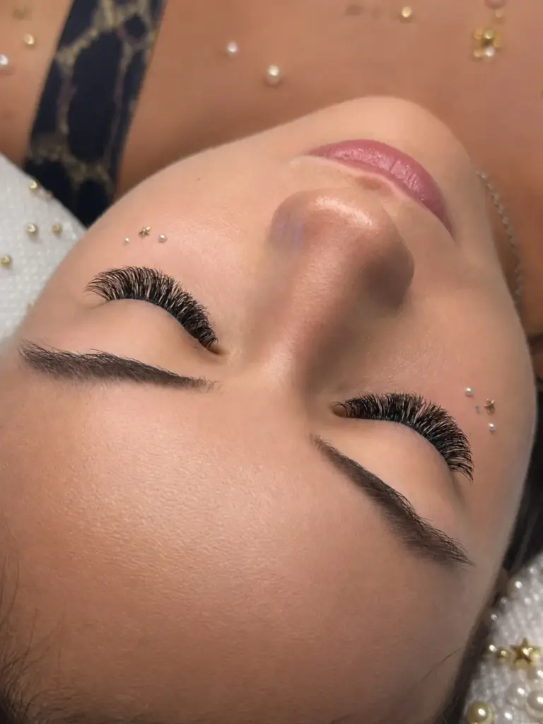 Close-up of a woman with eyes closed, long voluminous lash extensions, shaped brows, and decorative facial gems