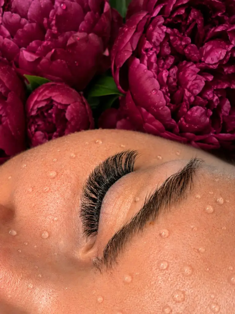 Close-up of a closed eye with long lash extensions and a shaped brow, framed by water droplets and burgundy flowers.