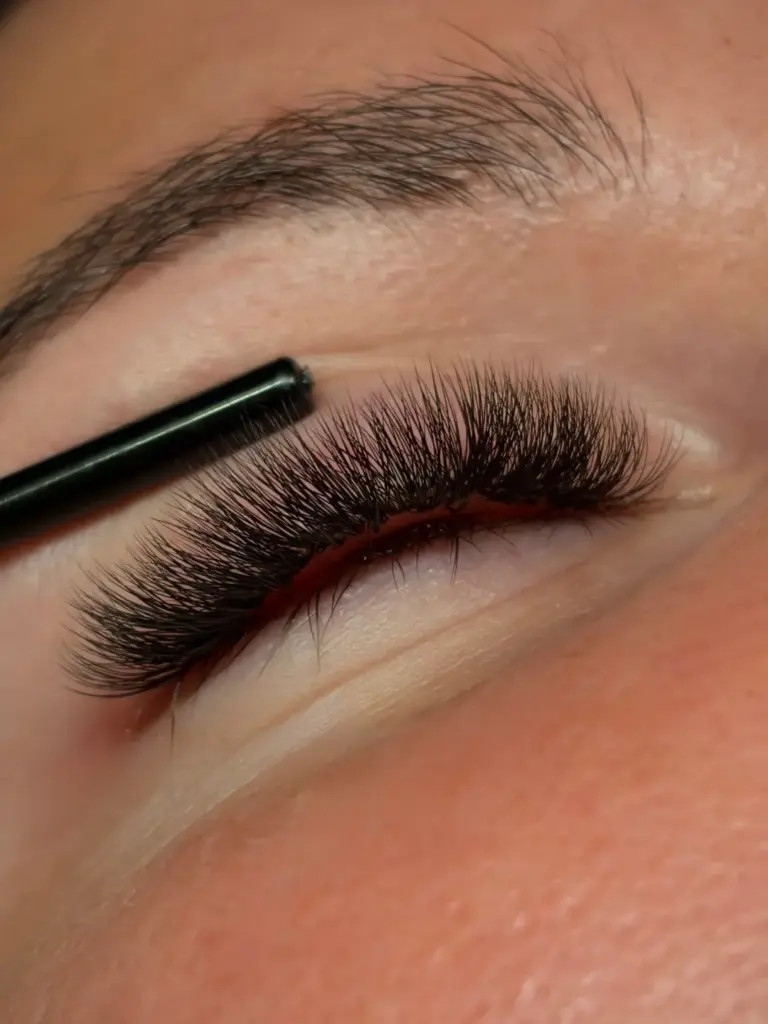 Close-up of dense, curled eyelash extensions being styled with a black tool beneath a natural eyebrow.